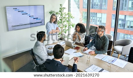 Senior female ceo and multicultural business people discussing company presentation at boardroom table. Diverse corporate team working together in modern meeting room office. Top view through glass