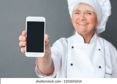 Senior Female Chef Studio Standing Isolated On Gray Wall Holding Smartphone Showign Screen Close-up Smiling Joyful Blurred Background