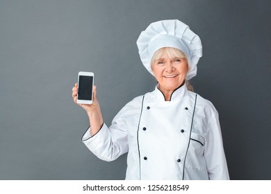 Senior Female Chef Studio Standing Isolated On Gray Wall Holding Smartphone Showign Empty Screen Looking Camera Smiling Cheerful