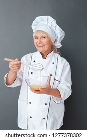 Senior Female Chef Studio Standing Isolated On Gray Wall Holding Whisk Mixing Eggs In Bowl Looking Camera Smiling Cheerful