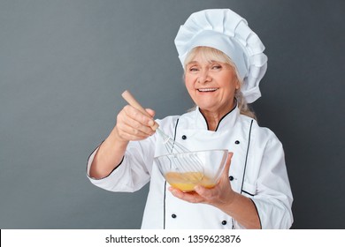 Senior Female Chef Studio Standing Isolated On Gray Wall Whisking Eggs In Bowl Cooking Omelette For Breakfast Looking Camera Smiling Happy Close-up