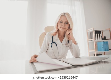 Senior female doctor reviewing medical records in a clinic office, highlighting dedication to healthcare and patient care. - Powered by Shutterstock