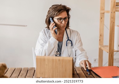 Senior female doctor talks on the phone while reviewing medical documents at her desk in the office. - Powered by Shutterstock