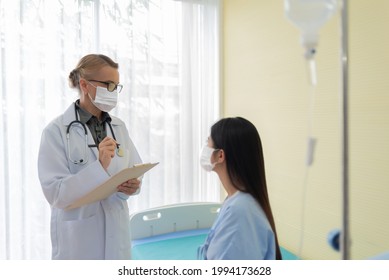 Senior Female Doctor Wearing Protective Face Mask Explaining Diagnosis To Her Asian Patient In Medical Room At Hospital.