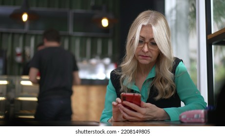 Senior Female Executive Woman Using Smartphone At Coffee Shop. Undecided Older Person Browsing Internet Online Seated At Cafe Place Restaurant