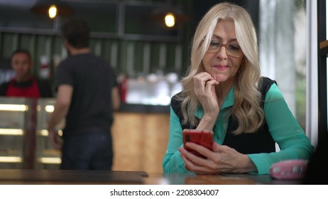 Senior Female Executive Woman Using Smartphone At Coffee Shop. Undecided Older Person Browsing Internet Online Seated At Cafe Place Restaurant