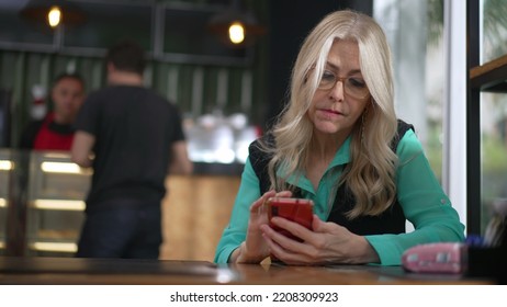 Senior Female Executive Woman Using Smartphone At Coffee Shop. Undecided Older Person Browsing Internet Online Seated At Cafe Place Restaurant