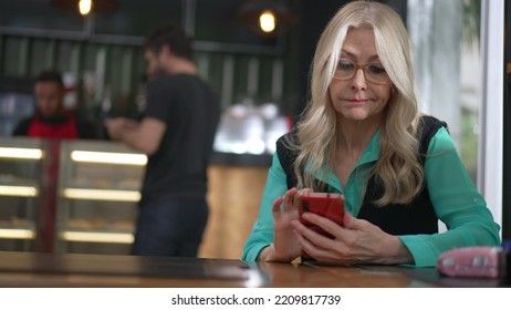 Senior Female Executive Woman Using Smartphone At Coffee Shop. Undecided Older Person Browsing Internet Online Seated At Cafe Place Restaurant