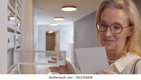 Senior Female In Eyewear Opening Postbox Standing In Hallway And Holding White Envelope With Invitation. Happy Mature Female Checking Mailbox And Smiling Receiving Good News
