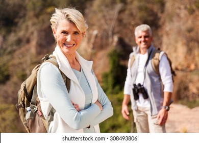 Senior Female Hiker Standing On Mountain With Husband On Background