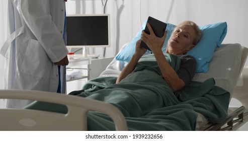 Senior Female Patient Lying In Hospital Bed And Signing Online Document On Digital Tablet. Doctor Giving Tablet Pc To Aged Sick Woman To Sign In Hospital Ward. Healthcare And Medicine Concept