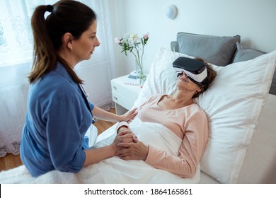 Senior Female Patient Relaxing In Hospital Bed with Virtual Reality Headset. Home Nurse Sitting on Bed Looking at Elderly Woman Getting Treatment Via VR Technology. - Powered by Shutterstock