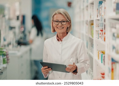 Senior female pharmacist is smiling while holding medication and using a digital tablet in a pharmacy with shelves full of medicine in the background - Powered by Shutterstock