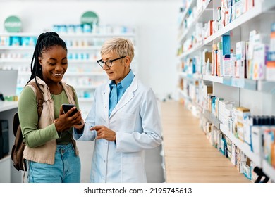 Senior Female Pharmacist And Young African American Customer Using Smart Phone In Drugstore.