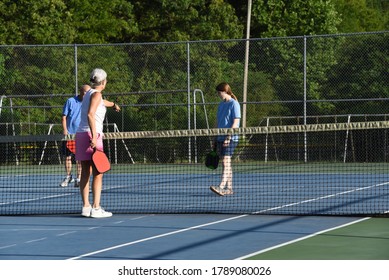 Senior Female Points And Explains The Rules Of Pickle Ball To A Young Female Player.  They Are Playing On An Outdoor Tennis Court.