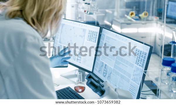 Senior Female Scientist Discusses Scientific\
Data with Her Laboratory Assistant. They\'re looking at Two Displays\
in a Modern Laboratory.