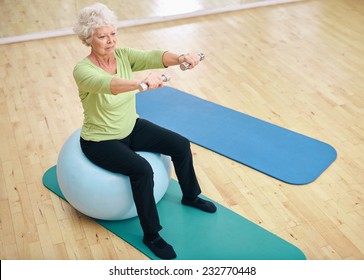 Senior Female Sitting On A Fitness Ball And Lifting Dumbbells. Old Woman Exercising With Weights At Gym.