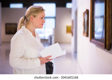 Senior Female Visitor Holding Guidebook Standing At Painting Exhibition In Art Museum