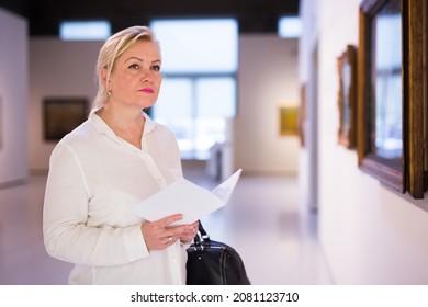 Senior Female Visitor Holding Guidebook Standing At Painting Exhibition In Art Museum