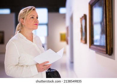 Senior Female Visitor Holding Guidebook Standing At Painting Exhibition In Art Museum