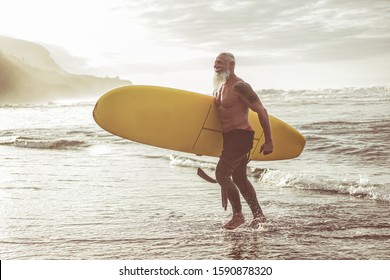 Senior fit guy walking with longboard after surfing at sunset - Mature tattooed man having fun doing extreme sport on tropical beach - Joyful elderly lifestyle and travel concept - Focus on male body - Powered by Shutterstock