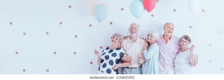 Senior Friends With Balloons Enjoying Retirement, Posing Together At A Birthday Party On A White Wall And Copy Space.