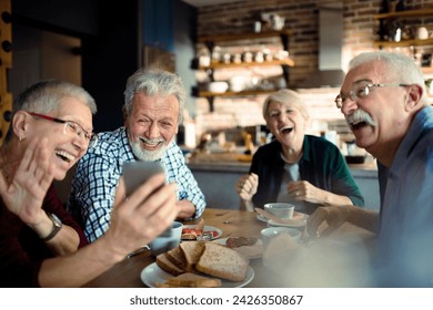 Senior friends laughing and sharing photos on smartphone at breakfast - Powered by Shutterstock