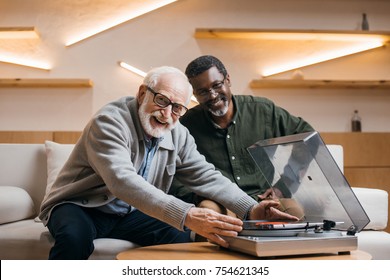 senior friends listening vinyl record while sitting on couch and looking at camera - Powered by Shutterstock