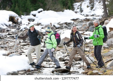 Senior Friends On Winter Vacation Hiking Over Stream In Snowy Forest