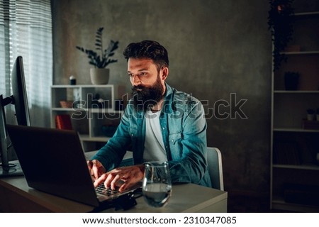 A senior full-stack developer is sitting in his darkened modern cozy home office and typing computer code on his laptop. A full-stack programmer is programming software at his home.
