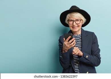 Senior Glad Woman Has Optical Eyewear Makes Banking Online On Cell Phone, Uses Modern Technologies For Searching Information In Internet, Dressed In Fashionable Apparel, Smiles Friendly, Isolated