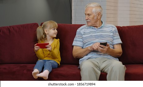 Senior grandfather with child girl granddaughter spending time home together, sitting on sofa in living room, using digital mobile phone. Watching videos, playing games, social network