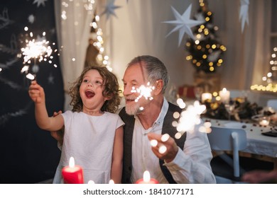 Senior grandfather with small granddaughter indoors at Christmas, sitting at table with sparklers.