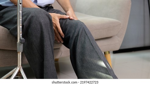 Senior Grandfather With Walking Stick Trying To Exercise Knee And Leg On Sofa At Home, Elderly Man With Knee Joint Pain And Suffering