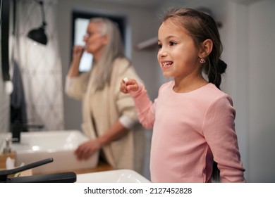 Senior grandmother and granddaughter standing indoors in bathroom, brushing teeth in morning.