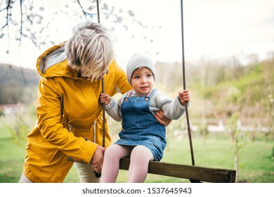 Senior grandmother with toddler granddaughter on a swing in garden in spring.