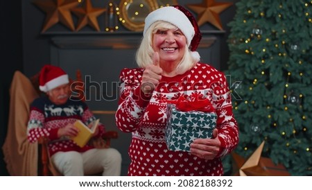 Senior grandmother woman in festive clothing at home with Christmas gift box smiling, raises thumbs up agrees with something or gives positive reply recommends advertisement likes good. Holidays mood