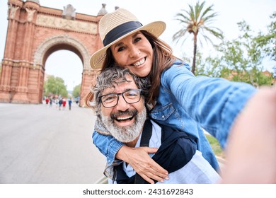 A senior gray hair man wearing a sun hat is giving a piggyback ride to a mature woman in a happy gesture while taking a selfie in touristic city. They seem to be having fun during their leisure travel - Powered by Shutterstock