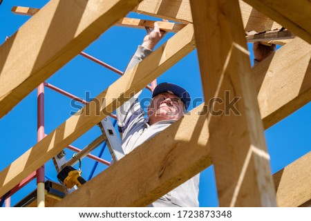 Senior gray-haired builder collects the frame of a wooden country house standing on the stairs against the blue sky. The physical activity of the elderly. Eco-friendly housing made of wood. Foto d'archivio © 