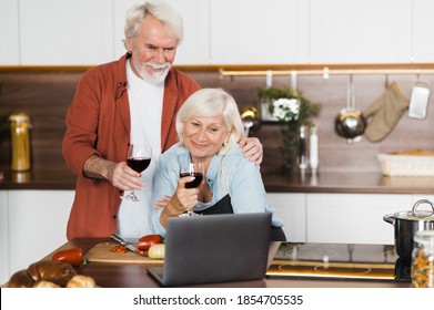Senior gray-haired married couple stand with a glass of wine in the kitchen, communicate by video call with family or friends using a laptop in the kitchen at home - Shutterstock ID 1854705535