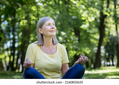 Senior gray-haired retired woman in the park meditates and performs breathing exercises, summer day in lotus position - Powered by Shutterstock