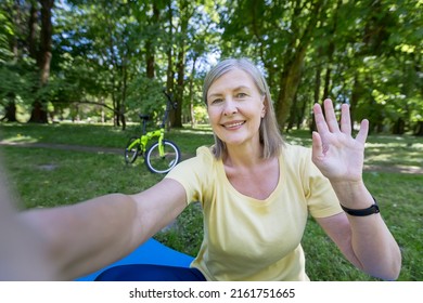 Senior gray-haired woman, active sportswoman in the park in the summer looks at the phone camera and smiles waving her hand at a video call