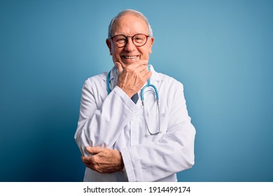 Senior grey haired doctor man wearing stethoscope and medical coat over blue background looking confident at the camera smiling with crossed arms and hand raised on chin. Thinking positive.