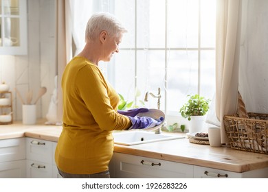 Senior Grey-haired Lady Washing Dishes In Cozy Kitchen, Using Rubber Gloves To Protect Her Hands, Back View, Copy Space. Attractive Grandmother Dishwashing At Home, Cleaning Plate With Cloth