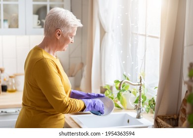 Senior Grey-haired Lady Washing Dishes In Cozy Kitchen, Using Rubber Gloves To Protect Her Hands, Back View, Copy Space. Attractive Grandmother Dishwashing At Home, Cleaning Plate With Cloth