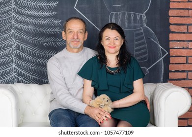 Senior Grey-haired Man Sitting On The White Sofa Hugging His Middle-aged Wife In Dark Green Dress. Happy Couple With Age Difference Looking At The Camera.