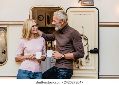 Senior grey-haired man touching his wife`s hair with love and talking in the motorhome doorway with cups of tea. Traveling caravanning together by trailer camper van - Powered by Shutterstock