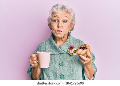 Senior Grey-haired Woman Drinking A Cup Of Coffee And Eating Bun Puffing Cheeks With Funny Face. Mouth Inflated With Air, Catching Air. 