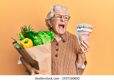 Senior grey-haired woman holding groceries and canadian dollars angry and mad screaming frustrated and furious, shouting with anger. rage and aggressive concept. 