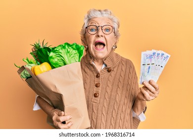Senior grey-haired woman holding groceries and colombian pesos banknotes celebrating crazy and amazed for success with open eyes screaming excited. 